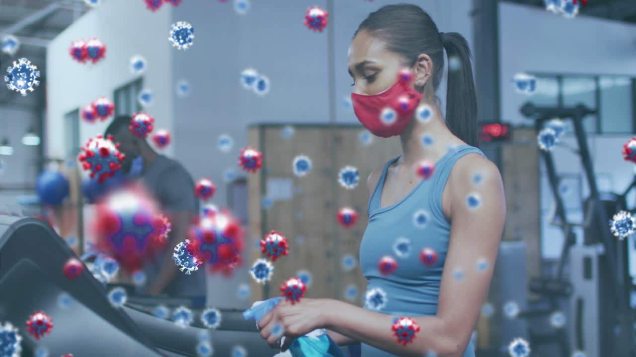 코로나19 세포가 마스크를 쓰고 체육관 장비를 소독제로 청소하는 여성에게 떠다니고 있습니다.