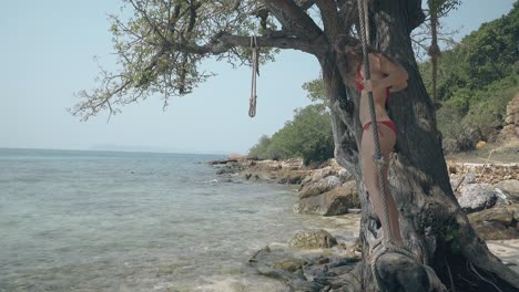 pretty woman in red bikini swings on tree log on ocean coast