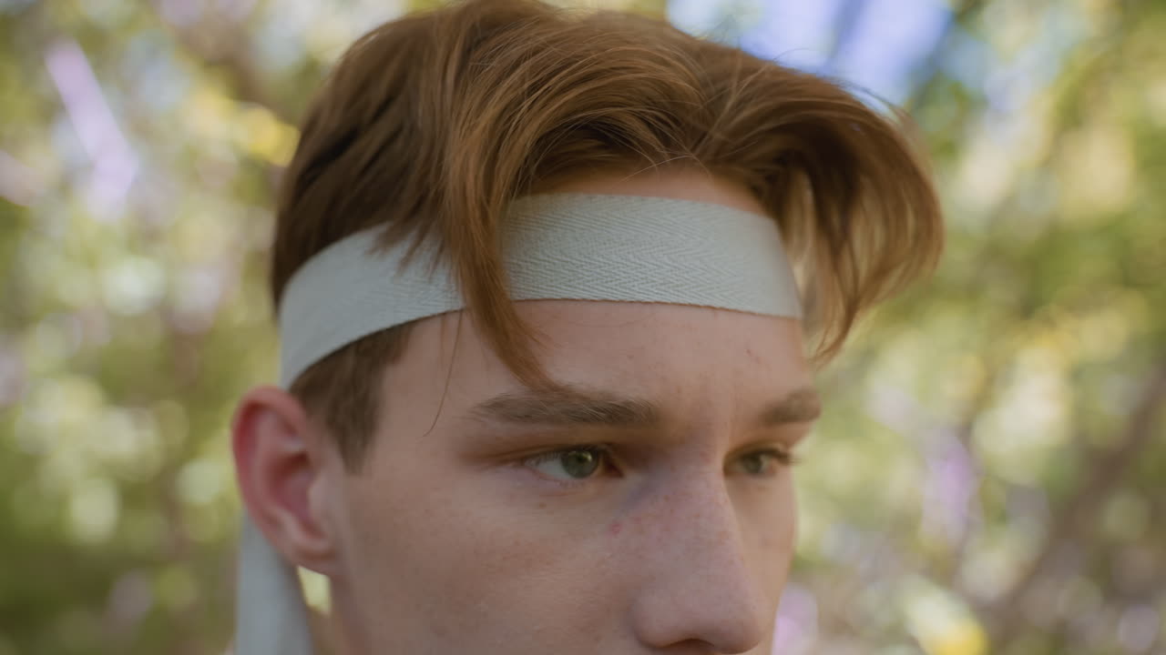 Close up view of young man winking while loosening white headband over forehead, freckles visible on skin, blurred woodland background in soft sunlight, playful yet focused expression