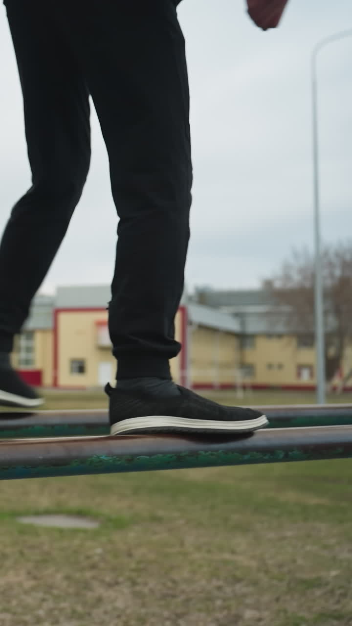 Close-up of a young boy carefully climbing on a rusted iron bar while balancing himself and walks on it, dressed in a black outfit and glasses, with a blurred background of trees and buildings