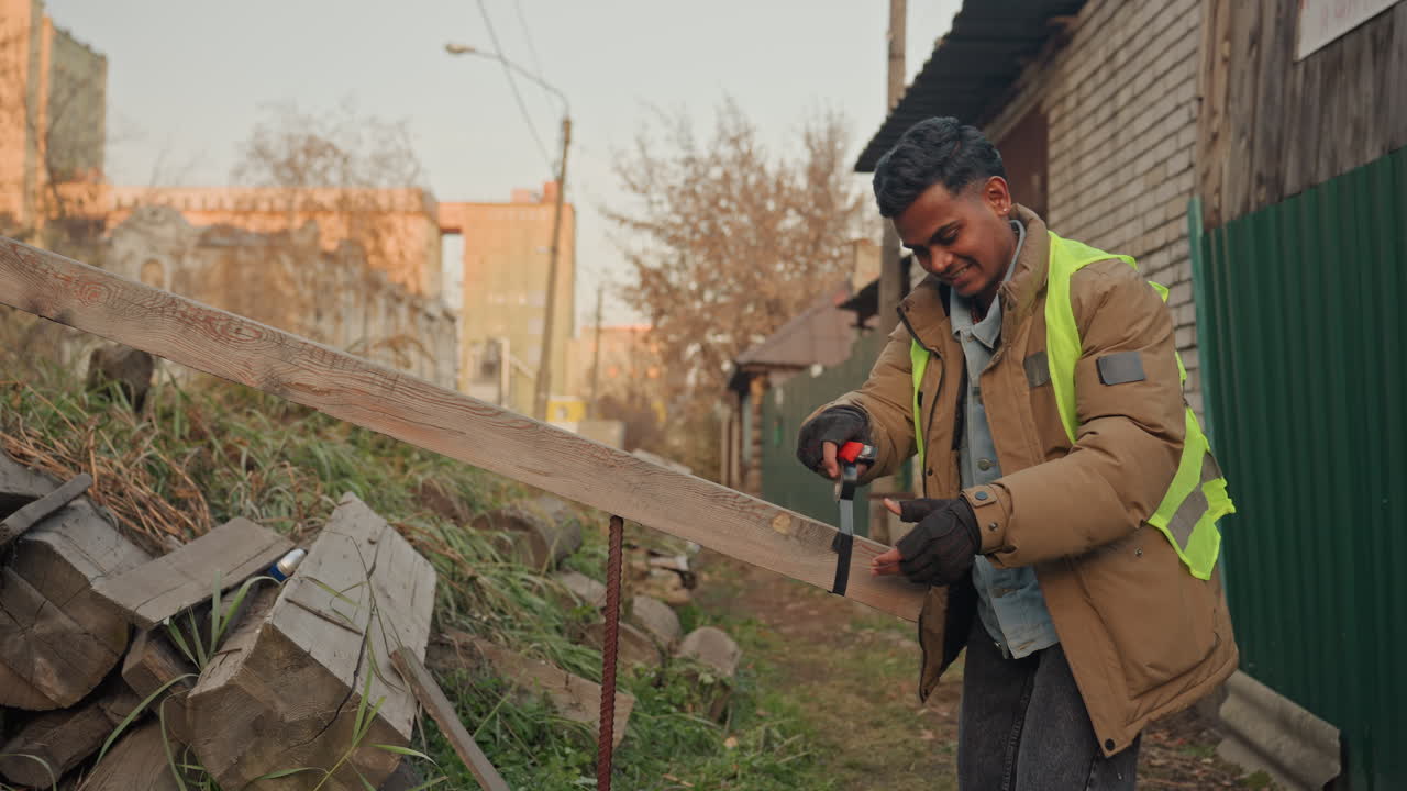 Street carpenter drilling wooden railing for repair, powering cordless driver to install screws, wearing reflective vest and gloves, leaning over stacked planks near backyard shed, active renovation