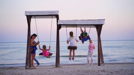 children having fun on wooden swings on the sea background. playground on sandy beach and children are swinging in the evening, looking at sea water.