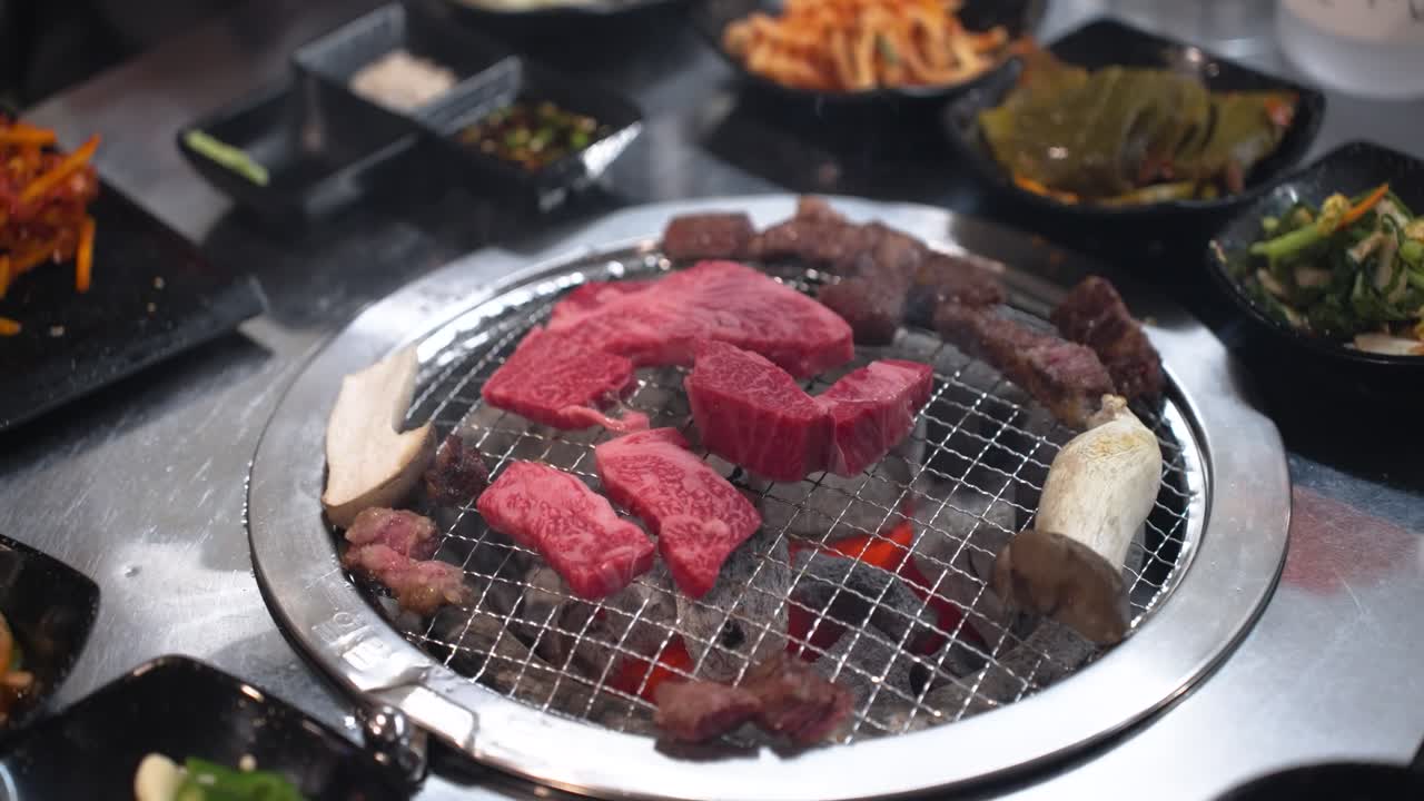 High-angle view of various cuts of raw, marbled Hanu beef cooking on a circular metal grill surrounded by small bowls of traditional Korean banchan on a black table