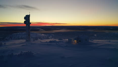 aerial view orbiting small winter hilltop cabin and transmission tower surrounded by snow covered landscape at sunset