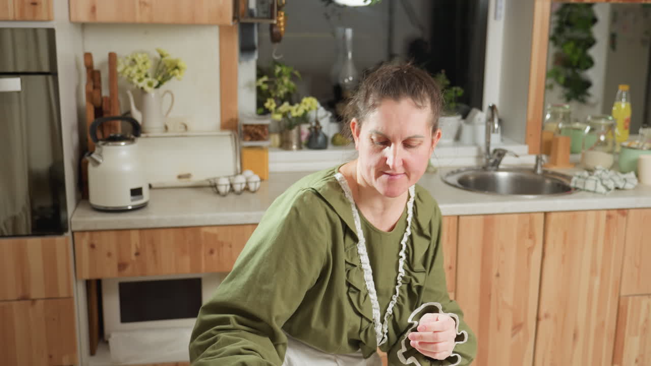 White woman dressed in green top and white apron drops white jug on kitchen table, then gently sits, places hand on face while gazing thoughtfully at items in front of her