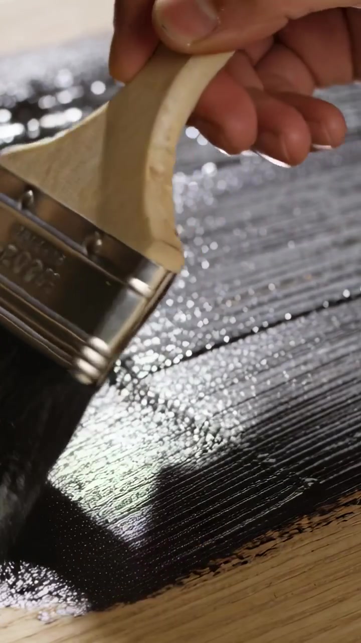 Vertical video: Loaded brush contacting plank, hand smoothing black paint, creating glossy finish