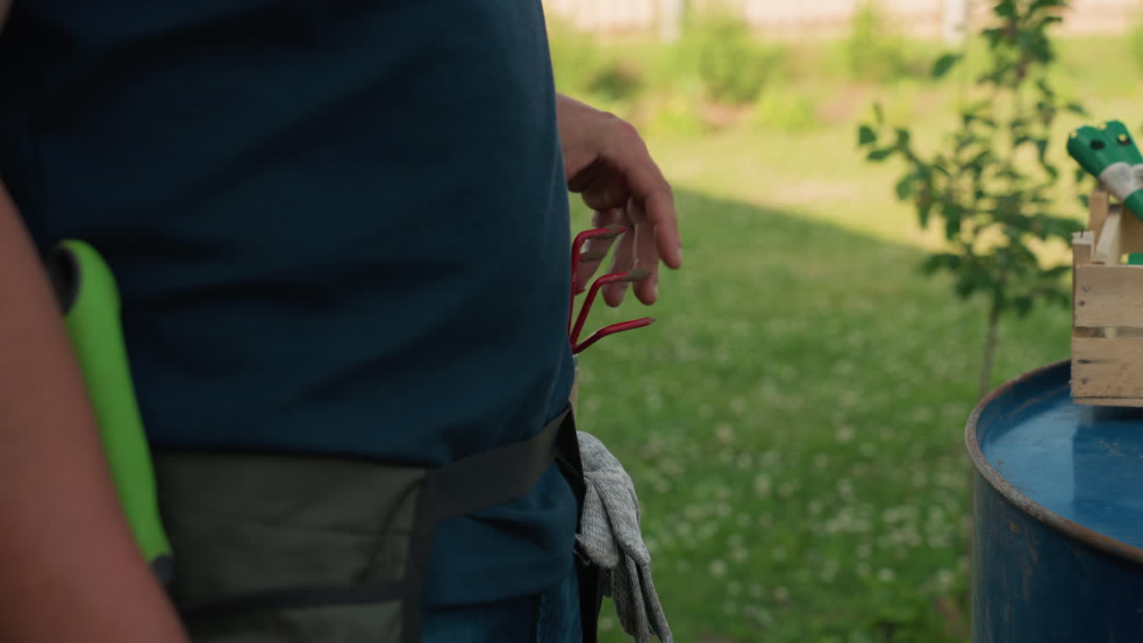 Male gardener wearing pocket apron picks red hand trowel fork and slides handle into side pocket beside wooden crate on blue drum, close view hands preparing tools for garden work