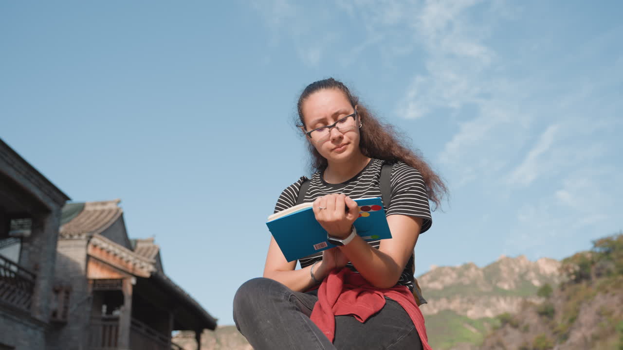 Giovane donna in un punto panoramico di montagna che scrive un diario su un quaderno sotto un cielo sereno, collina verde e cime distanti, postura tranquilla e riflessiva con lo zaino, la luce del sole illumina capelli e occhiali, serena.