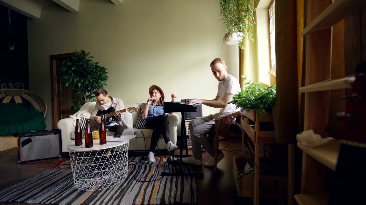 Attractive young woman vocalist is singing in microphone while her friends guys are playing the keyboard and the guitar in spacious light home studio.