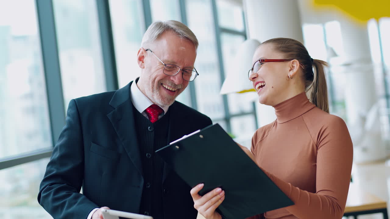 Young woman assistant and senior businessman working together in a light room with big windows, laughing. Team work. Business video