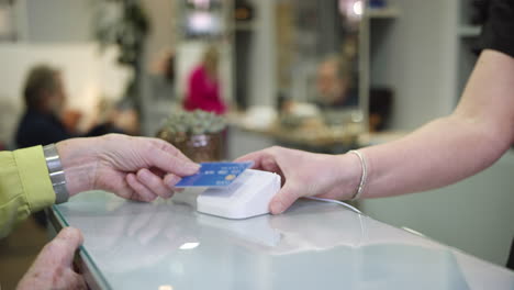 close up of senior woman making contactless payment to stylist in salon with credit card