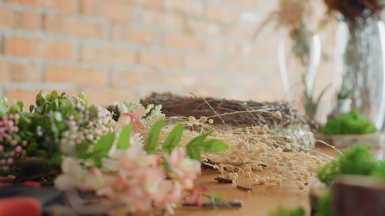 Close view of flowers, greenery, and dried stems on wooden table near unfinished wreath base prepared for handmade floral decoration project with natural materials inside rustic indoor creative workshop
