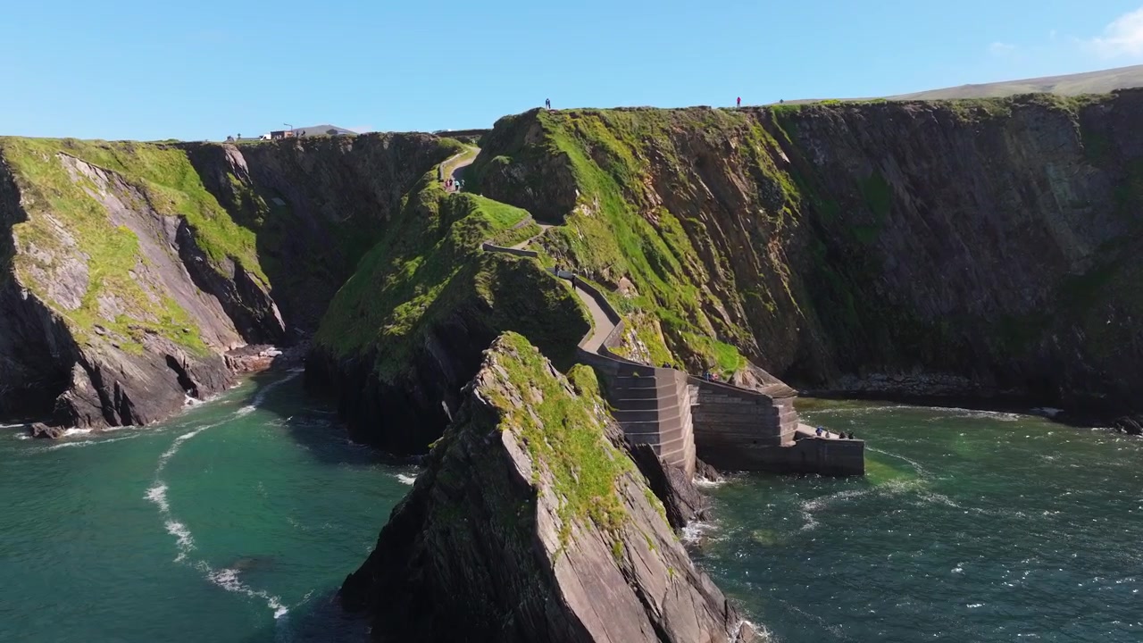 A winding pathway traces the edge of steep cliffs at Dunquin Pier, with clear blue waters below. Perfect for travel features or Irish landscape documentaries.