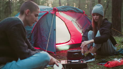 woman in beanie and tracksuit seated near tent hands over canned food to man as pot boils on portable gas stove during camping activity surrounded by cooking supplies and forest trees