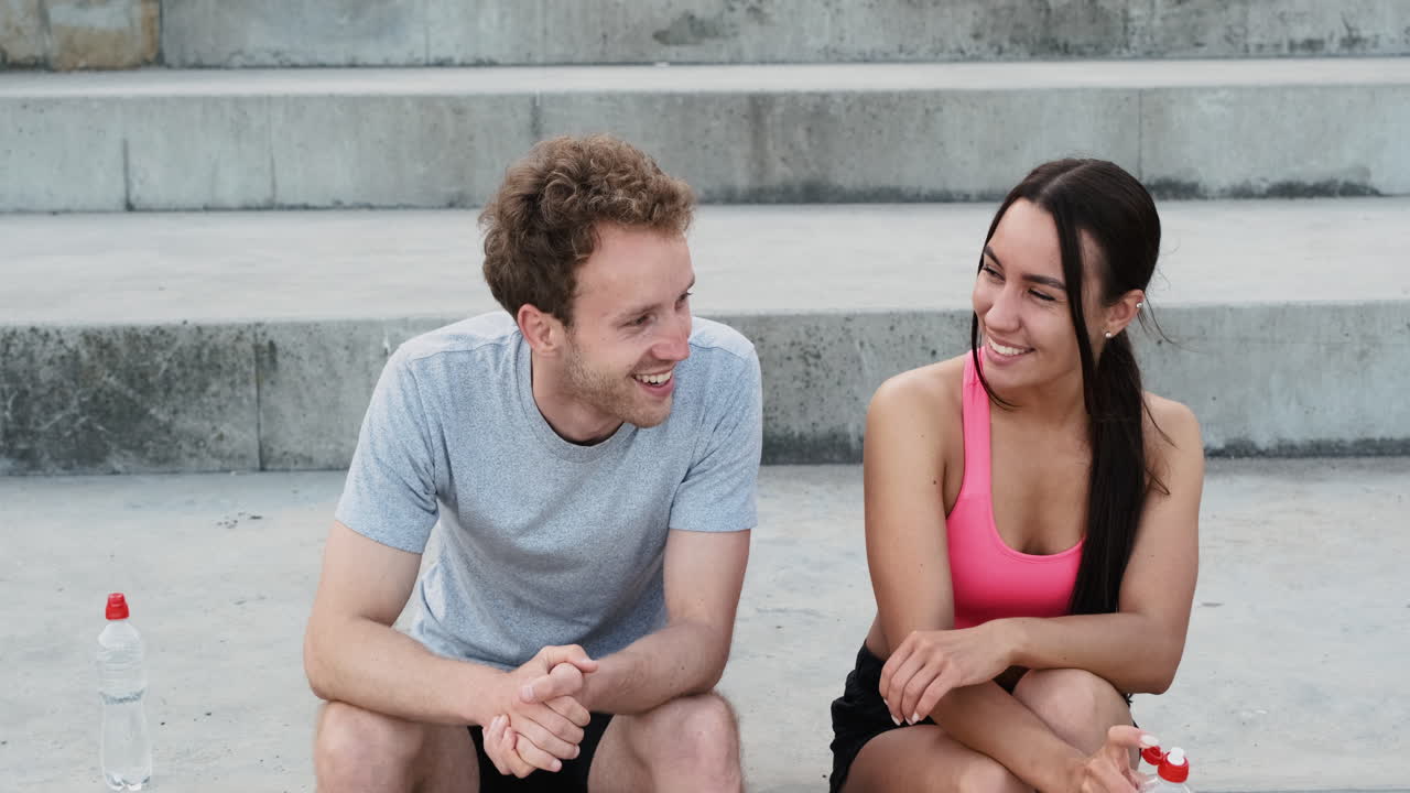 feliz pareja deportiva sentada en las escaleras y hablando entre ellos después de una sesión en la ciudad