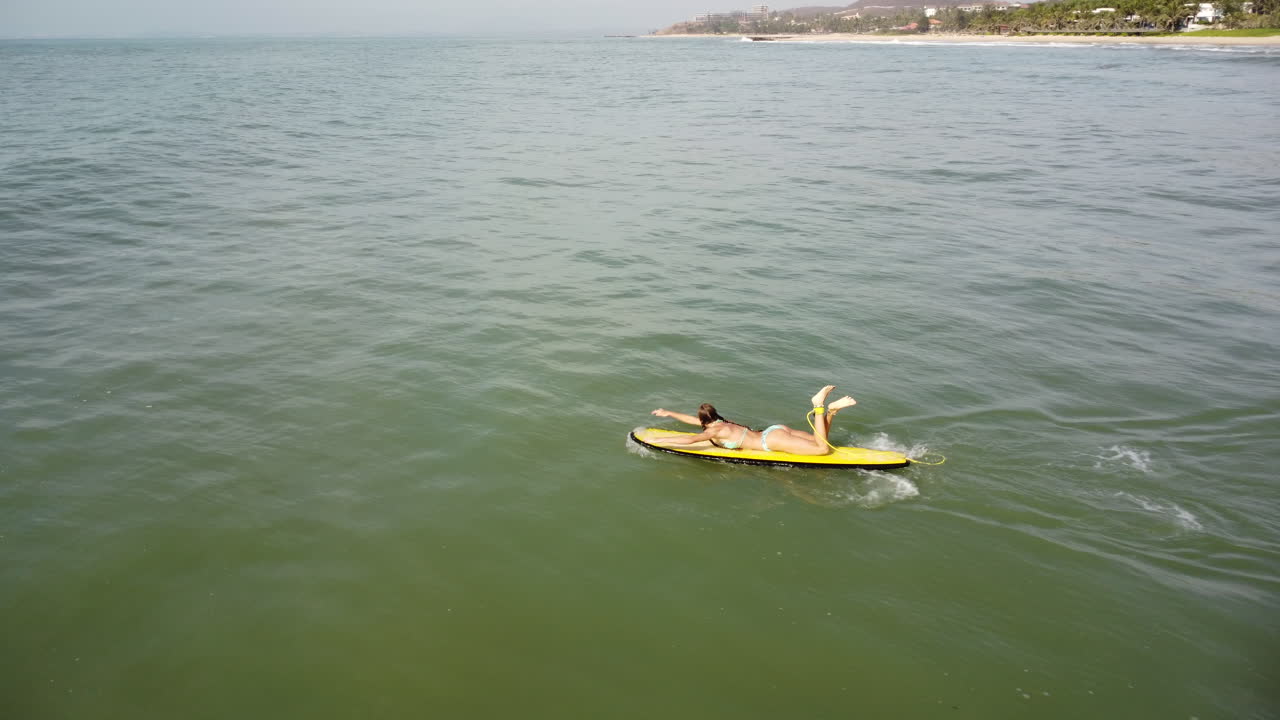 joven surfista sexy caucásica en bikini remando en una tabla de surf amarilla en agua de mar limpia y abierta durante un día soleado de vacaciones de verano