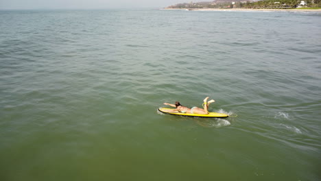 Caucasian-young-sexy-surfer-in-bikini-paddling-on-yellow-surf-board-in-open-clean-ocean-water-during-a-sunny-day-of-summer-holiday