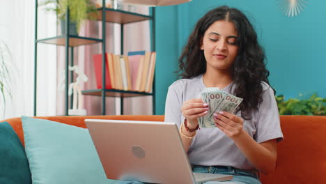 Una-Mujer-Feliz-Y-Sonriente-Contando-Dinero-En-Efectivo-Y-Usando-Una-Computadora-Portátil-Para-Calcular-Los-Ingresos-Domésticos-En-Casa