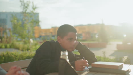 Kid writing on wooden table with notebook and pen while woman prepares to bring chips from bag, showing balance of study, care, and snack time in bright outdoor park environment