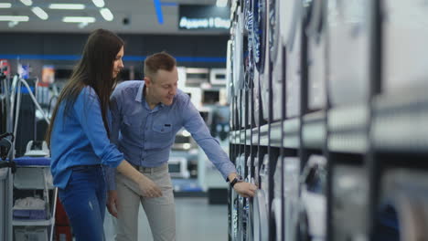 young married couple man and woman in casual clothes appliances store choose to buy washing machine for home. open the door looking into the drum, compare the design and characteristics of the devices.