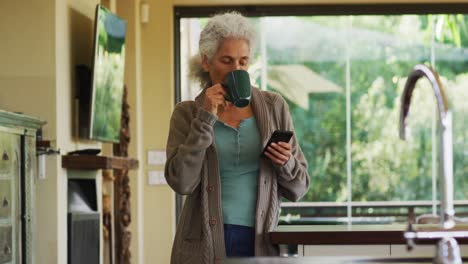 Portrait-of-senior-mixed-race-woman-drinking-coffee-and-using-smartphone-in-kitchen