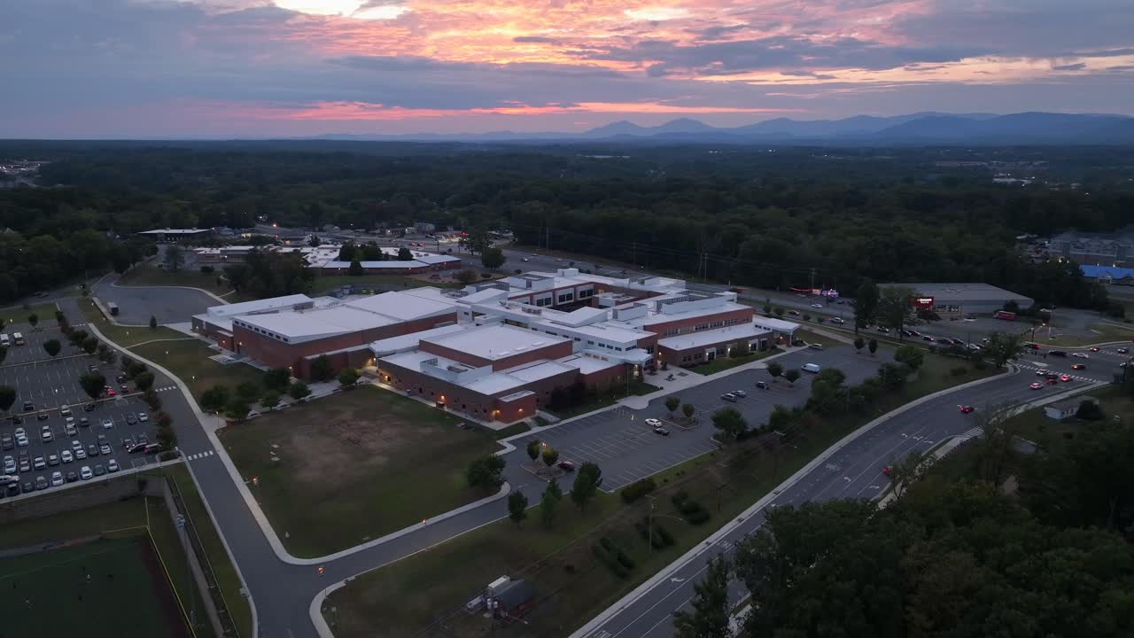 Dusk scene in suburb of American town. American school in red brick design and traffic on highway road. Aerial rising wide shot. Colored sky in late summer season. Panorama view
