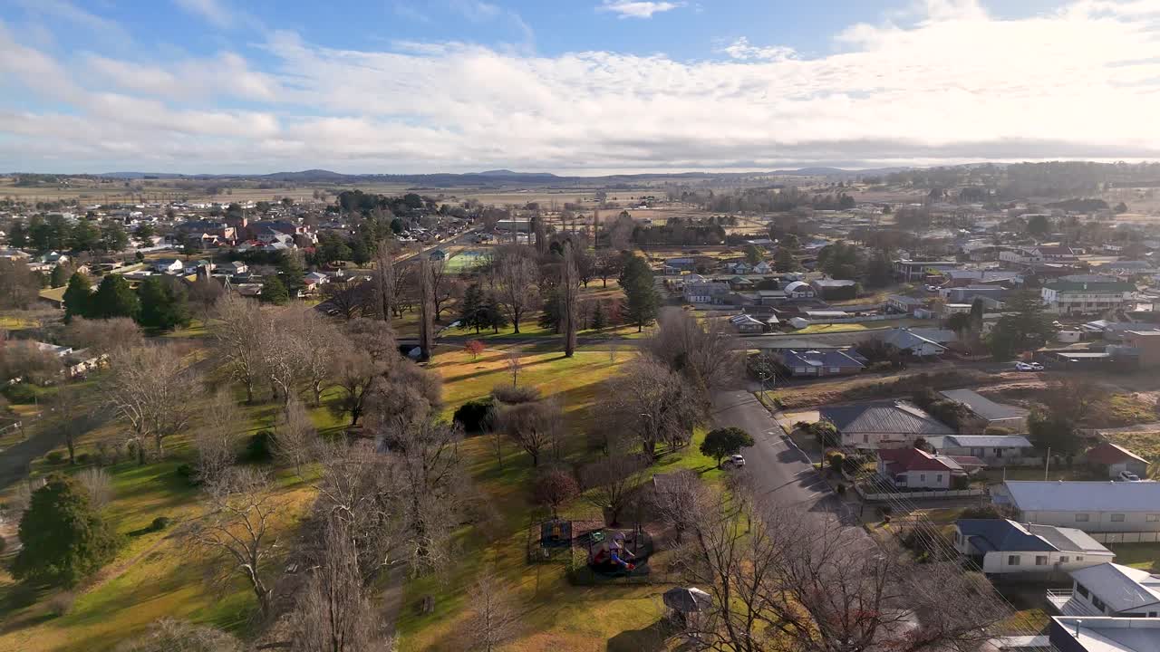 Drone glides above a public park with leafless trees in Coonabarabran, New South Wales, under soft daylight, revealing residential streets and distant hills
