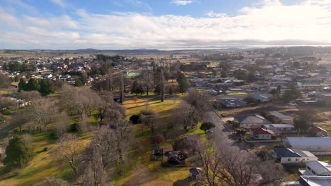 drone glides above a public park with leafless trees in coonabarabran, new south wales, under soft daylight, revealing residential streets and distant hills
