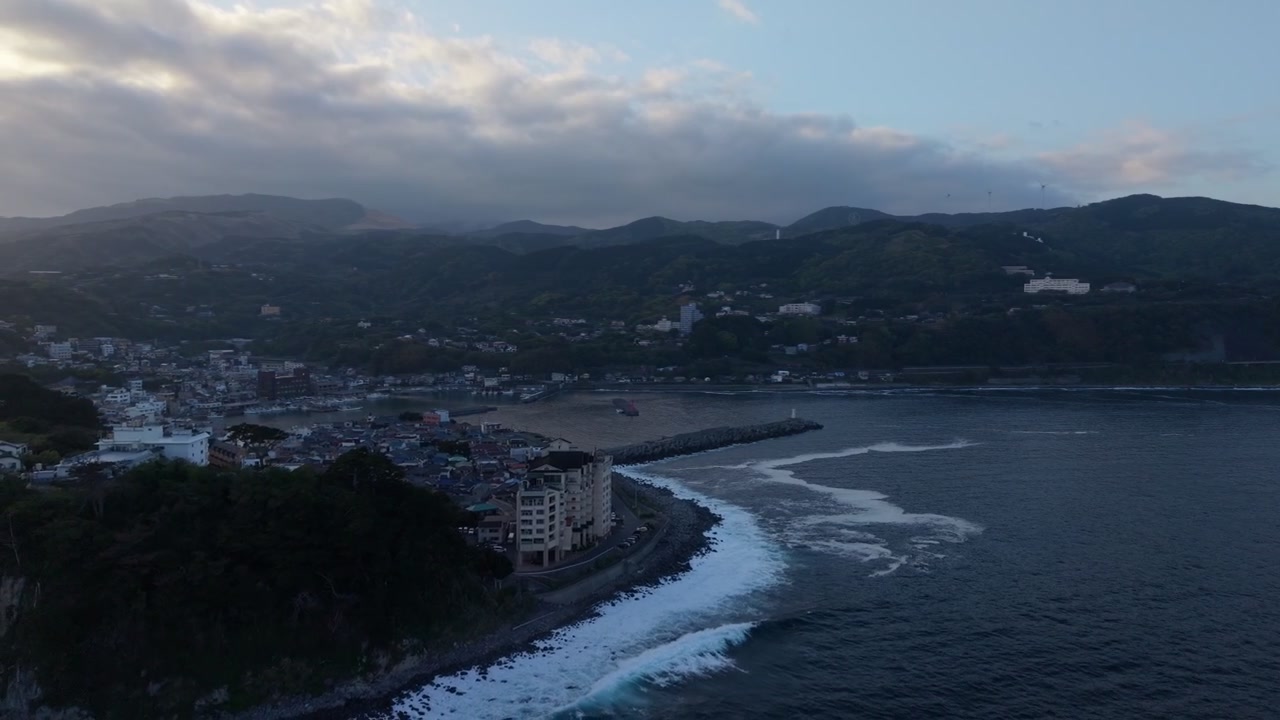 Cittadina portuale con un lungo frangiflutti in pietra e un piccolo faro sul molo che si curva nel mare a Higashiizu, Izu, Giappone, in un pomeriggio nuvoloso, ripresa aerea con drone