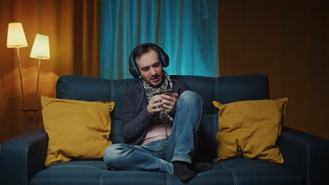 hombre caucásico con auriculares escuchando música en el teléfono inteligente a través de auriculares en la sala de estar.