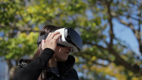 young dark-haired woman in warm black waistcoat wearing virtual reality glasses in early autumn park