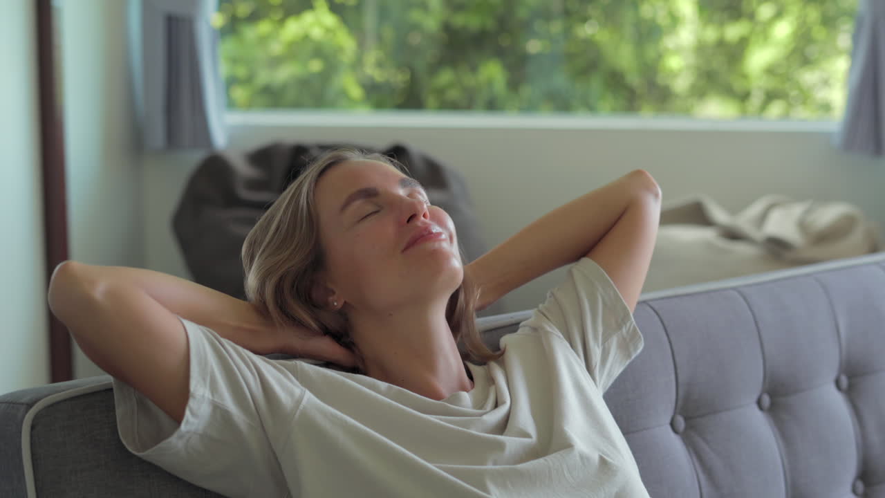 Yong woman closing hands behind hand breathing fresh air in living room resting in comfortable sofa. Calm young woman dozing daytime nap on couch with closed eyes