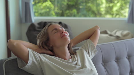 yong woman closing hands behind hand breathing fresh air in living room resting in comfortable sofa. calm young woman dozing daytime nap on couch with closed eyes