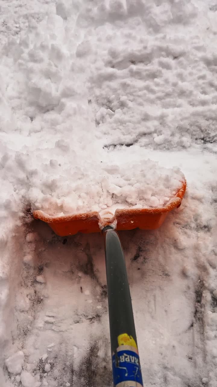 POV de uma pá de neve raspando uma calçada residencial em pequenos empurrões, revelando gelo negro por baixo. Formato vertical.