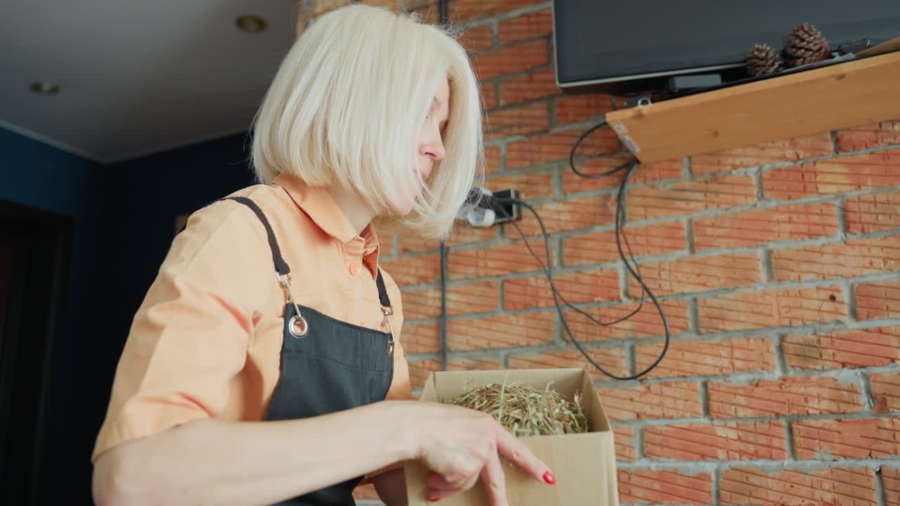 Florist in apron with blonde hair holding cardboard box filled with hay and wooden packaging inside workshop with brick wall background, preparing eco friendly container for handmade craft