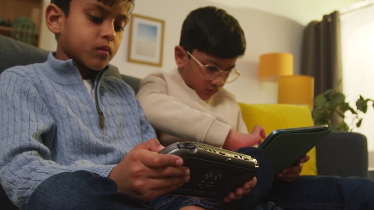 Due Giovani Ragazzi Seduti sul Divano a Casa Giocano o Guardano Contenuti in Streaming su un Tablet Digitale e un Dispositivo di Gioco Portatile