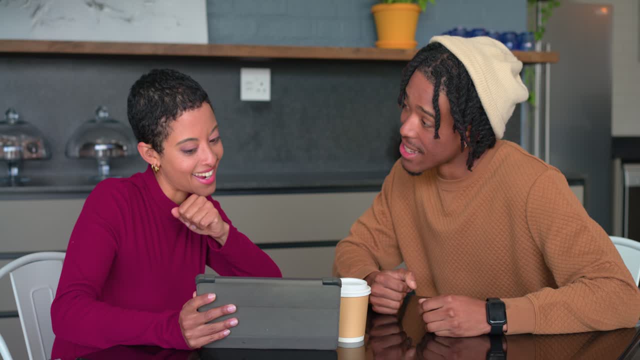 Diverse couple placing pastry then tapping tablet while sharing recipe ideas at kitchen table