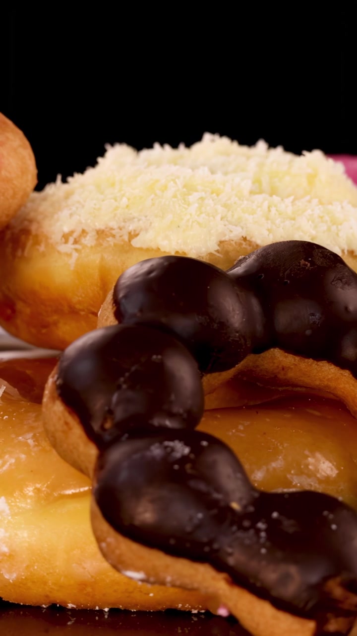 Primo piano rotante di ciambelle assortite con glassa al cioccolato, alla fragola e zucchero a velo, su uno sfondo nero con illuminazione brillante e uniforme