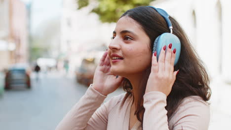 Happy-Indian-young-woman-wearing-wireless-headphones-listening-music-dancing-on-city-street-outdoors