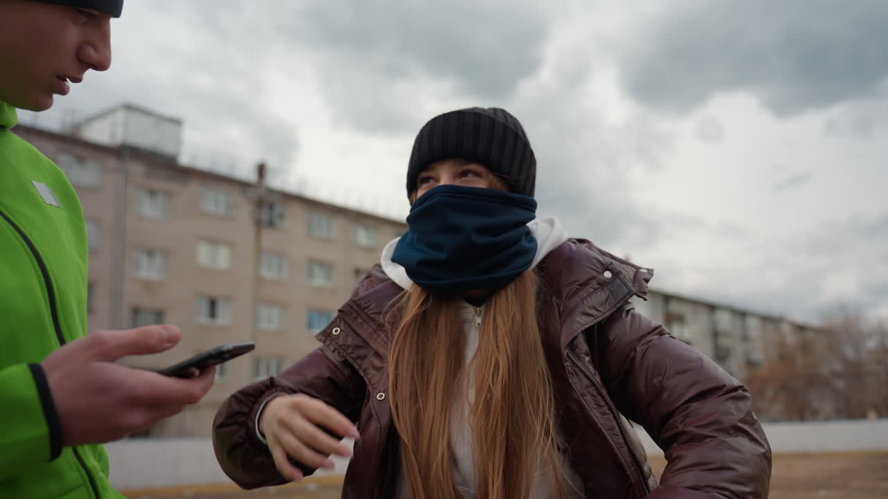 차가운 바람이 부는 흐린 날씨에 녹색 재킷, 비니, 갈색 코트를 입은 배달원과 백인 여성이 긴장된 분위기 속에서 길거리에서 대화를 나누고 있으며, 여성은 얼굴에 스카프를 두르고 있고, 배경에는 아파트 건물이 보입니다.