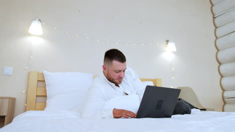calm caucasian man lying on the bed looks at his laptop. man uses gadget for work remote or fun.