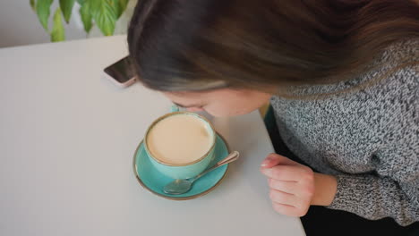 aerial view of young woman in knit sweater smelling hot tea in green cup with saucer and spoon on table near smartphone cozy indoor setting with soft lighting and plant in background calm