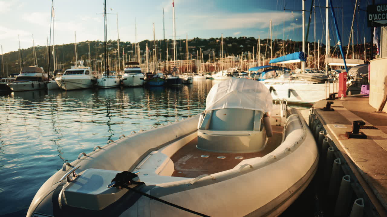 Un motoscafo gonfiabile ormeggiato a una banchina in un trafficato porticciolo turistico, con barche a vela e yacht che si riflettono nell'acqua calma sotto la luce soffusa del pomeriggio
