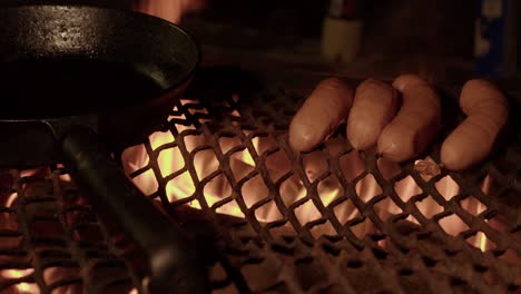 las salchichas se sientan al lado de una sartén de hierro en una parrilla de metal sobre un fuego abierto por la noche
