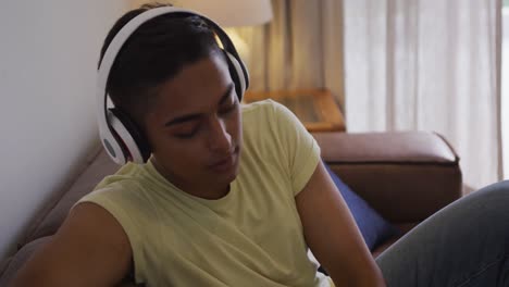 Mixed-race-man-wearing-headphones-sitting-on-sofa-using-smartphone-and-laughing