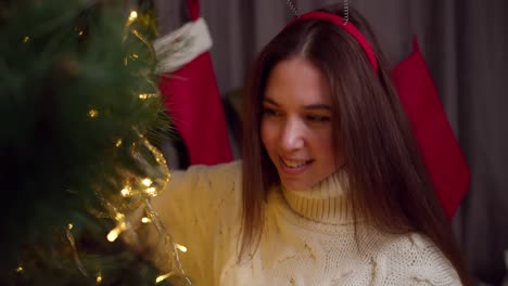 Close-up-shot-of-a-happy-brunette-girl-in-a-white-sweater-decorates-the-New-Year-tree-with-the-help-of-a-yellow-glowing-garland-in-a-cozy-home-in-winter-and-prepares-for-the-New-Year-holidays-and-Christmas