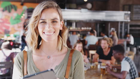 portrait of waitress holding digital tablet in busy bar restaurant smiling at camera