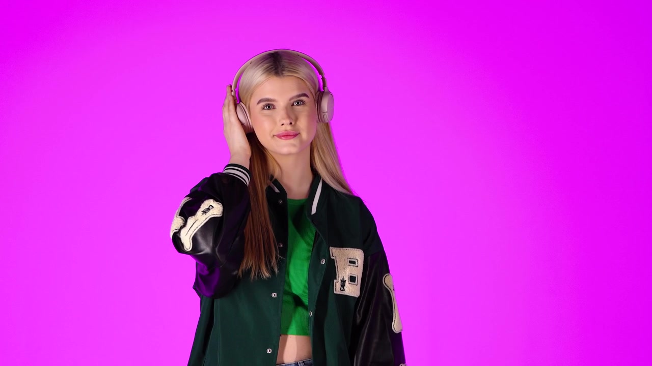 Bella giovane donna bionda che ascolta musica con le cuffie e balla, ritratto in studio con sfondo rosa.