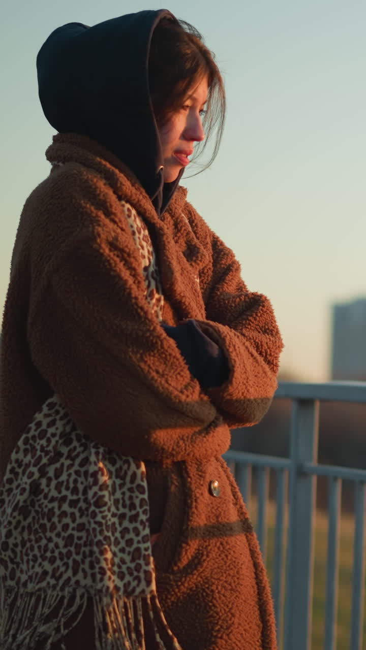 una chica melancólica está cerca de una barandilla de metal, envuelta en un abrigo marrón cálido y una sudadera con capucha. mira hacia la distancia, perdida en sus pensamientos