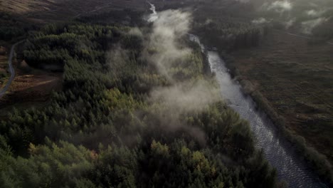 aerial drone footage slowly reversing and tilting to reveal a river, road and forest of conifer trees while low hanging cloud hugs the treetops and the sun sets behind mountains